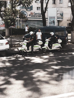 Two individuals wearing helmets and riding scooters are parked on the side of a street. They appear to be part of a law enforcement or security service, as indicated by the markings on their scooters. There are two men standing nearby, one using a phone and another observing the scene. In the background, several buildings feature business signage, and trees provide some shade on the street.