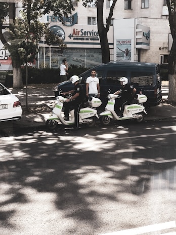 Two individuals wearing helmets and riding scooters are parked on the side of a street. They appear to be part of a law enforcement or security service, as indicated by the markings on their scooters. There are two men standing nearby, one using a phone and another observing the scene. In the background, several buildings feature business signage, and trees provide some shade on the street.