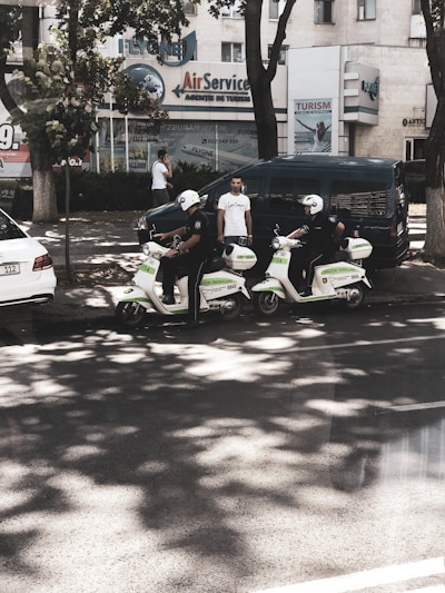 Two individuals wearing helmets and riding scooters are parked on the side of a street. They appear to be part of a law enforcement or security service, as indicated by the markings on their scooters. There are two men standing nearby, one using a phone and another observing the scene. In the background, several buildings feature business signage, and trees provide some shade on the street.
