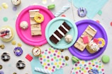 An assortment of colorful, decorated desserts is displayed on vibrant paper plates. There are slices of cake, cupcakes with frosting and sprinkles, and miniature donuts. The scene is set against a backdrop of pastel napkins and festive confetti scattered around the surface.