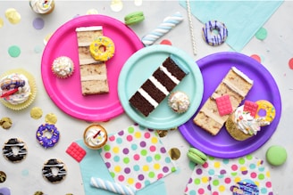 An assortment of colorful, decorated desserts is displayed on vibrant paper plates. There are slices of cake, cupcakes with frosting and sprinkles, and miniature donuts. The scene is set against a backdrop of pastel napkins and festive confetti scattered around the surface.