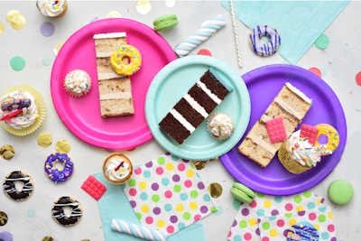 An assortment of colorful, decorated desserts is displayed on vibrant paper plates. There are slices of cake, cupcakes with frosting and sprinkles, and miniature donuts. The scene is set against a backdrop of pastel napkins and festive confetti scattered around the surface.