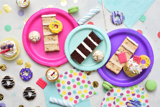 An assortment of colorful, decorated desserts is displayed on vibrant paper plates. There are slices of cake, cupcakes with frosting and sprinkles, and miniature donuts. The scene is set against a backdrop of pastel napkins and festive confetti scattered around the surface.