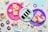 An assortment of colorful, decorated desserts is displayed on vibrant paper plates. There are slices of cake, cupcakes with frosting and sprinkles, and miniature donuts. The scene is set against a backdrop of pastel napkins and festive confetti scattered around the surface.