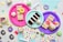 An assortment of colorful, decorated desserts is displayed on vibrant paper plates. There are slices of cake, cupcakes with frosting and sprinkles, and miniature donuts. The scene is set against a backdrop of pastel napkins and festive confetti scattered around the surface.