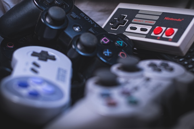 A collection of vintage and modern video game controllers includes devices from different gaming consoles. The controllers feature various buttons and joysticks, with some showcasing iconic buttons like the red and white buttons on an old Nintendo controller.