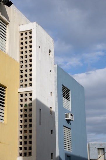 A building with a modern architectural design featuring yellow, white, and blue sections. The building has rectangular windows with shutters and an external air conditioning unit. The white section has a unique design with hollow square patterns, and shadows are cast across the structure.