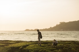 Two people are exploring a rocky, moss-covered shore near the ocean. One person is standing, holding a stick or similar object, while the other is crouched down, possibly examining the ground. The ocean is calm, with gentle waves, and there is a hilly landscape in the background. The lighting suggests it is either early morning or late afternoon, casting a warm golden hue over the scene.