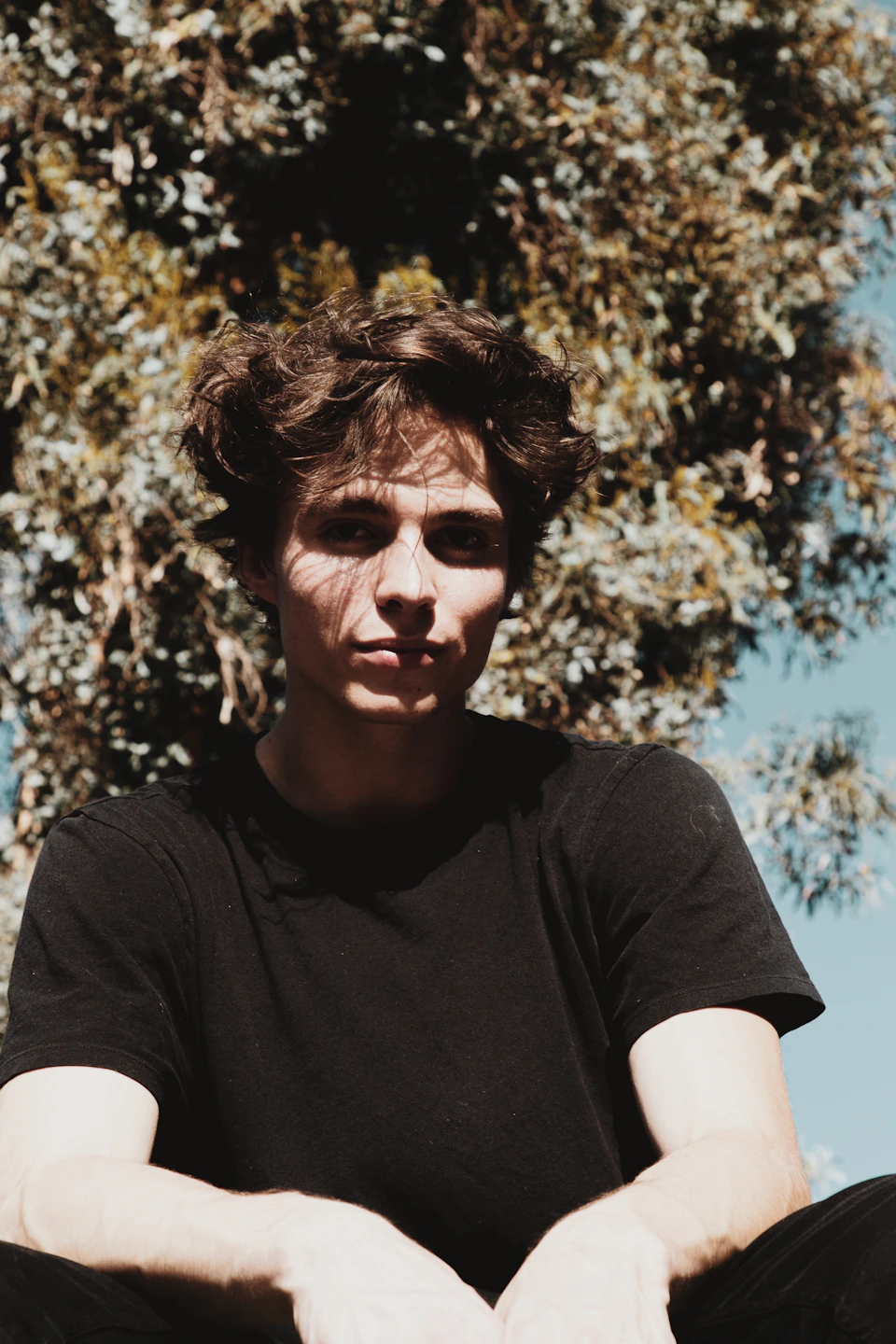 The image shows a young man with curly hair sitting under a tree, dressed in a black t-shirt and pants. The tree's leaves are green and lush against the clear blue sky, creating a serene backdrop for the scene. The man is positioned on the left side of the frame, facing towards the right, giving a sense of depth to the image.