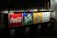 A brightly illuminated billboard displaying logos for Coca-Cola, Sprite, and Fanta. Each logo is prominently displayed against distinct colored backgrounds: red for Coca-Cola, green for Sprite, and orange for Fanta. To the right, there is text in Chinese. The surrounding environment appears dark and urban.