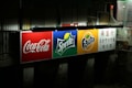 A brightly illuminated billboard displaying logos for Coca-Cola, Sprite, and Fanta. Each logo is prominently displayed against distinct colored backgrounds: red for Coca-Cola, green for Sprite, and orange for Fanta. To the right, there is text in Chinese. The surrounding environment appears dark and urban.