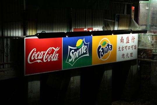 A brightly illuminated billboard displaying logos for Coca-Cola, Sprite, and Fanta. Each logo is prominently displayed against distinct colored backgrounds: red for Coca-Cola, green for Sprite, and orange for Fanta. To the right, there is text in Chinese. The surrounding environment appears dark and urban.