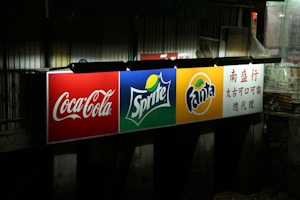 A brightly illuminated billboard displaying logos for Coca-Cola, Sprite, and Fanta. Each logo is prominently displayed against distinct colored backgrounds: red for Coca-Cola, green for Sprite, and orange for Fanta. To the right, there is text in Chinese. The surrounding environment appears dark and urban.