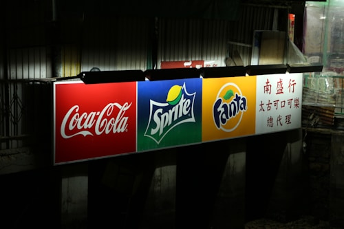 A brightly illuminated billboard displaying logos for Coca-Cola, Sprite, and Fanta. Each logo is prominently displayed against distinct colored backgrounds: red for Coca-Cola, green for Sprite, and orange for Fanta. To the right, there is text in Chinese. The surrounding environment appears dark and urban.