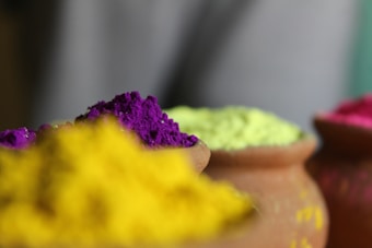 Brightly colored powders in clay pots arranged closely, featuring vibrant shades of yellow, purple, green, and pink. The focus is on the textures and vividness of the colors, with a blurred background.