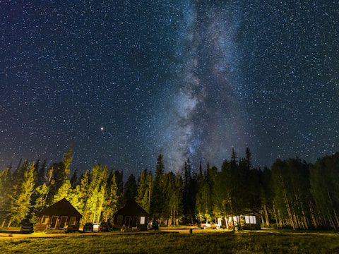 A breathtaking night sky filled with innumerable stars and the Milky Way galaxy stretching across the sky. Below, a serene forest setting with several small, illuminated cabins nestled among tall trees. Cars are parked beside the cabins, adding to the peaceful and secluded atmosphere.