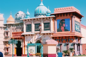 A vibrant architectural scene featuring a building with ornate decorations and domed rooftops, inspired by traditional Indian design. Prominent billboards display posters from classic Indian movies, adding a cinematic atmosphere. The area includes colorful signs and banners, evoking a festive and cultural ambiance.