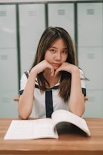 A young woman in a sailor-style uniform sits at a wooden desk with an open book in front of her. She has a focused expression, resting her chin on her hands, with lockers visible in the background.