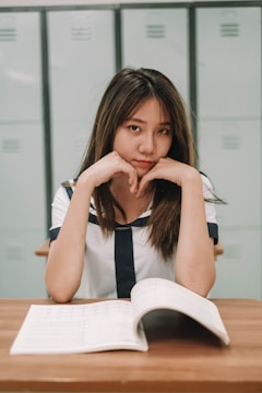 A young woman in a sailor-style uniform sits at a wooden desk with an open book in front of her. She has a focused expression, resting her chin on her hands, with lockers visible in the background.