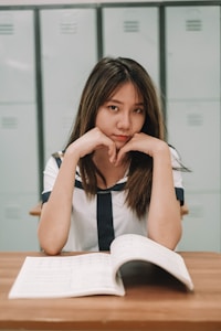 A young woman in a sailor-style uniform sits at a wooden desk with an open book in front of her. She has a focused expression, resting her chin on her hands, with lockers visible in the background.