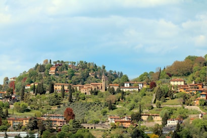 A scenic hillside landscape featuring a collection of picturesque houses and villas surrounded by lush greenery and trees. The structures are scattered across the gentle slopes, nestled among dense foliage, creating a tranquil and charming view of the countryside. The sky above is a bright, clear blue with some fluffy clouds adding depth to the scene.