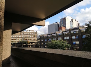 A broad view of modern and brutalist architecture in an urban environment. The structures have a mix of concrete and glass materials, with several windows displaying reflections. A clear blue sky with some clouds forms the backdrop, highlighting the rigid lines and angles of the buildings. Greenery can be seen in the form of trees and plants on balconies and terraces.