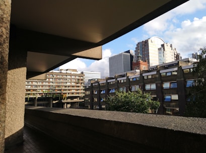 A broad view of modern and brutalist architecture in an urban environment. The structures have a mix of concrete and glass materials, with several windows displaying reflections. A clear blue sky with some clouds forms the backdrop, highlighting the rigid lines and angles of the buildings. Greenery can be seen in the form of trees and plants on balconies and terraces.