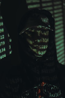 A person in a hoodie and cap is partially illuminated by a digital light projection of numbers, creating a mysterious and tech-oriented atmosphere. The background is dark, enhancing the focus on the numerical overlay across the person's face and clothing.