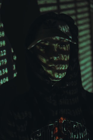 A person in a hoodie and cap is partially illuminated by a digital light projection of numbers, creating a mysterious and tech-oriented atmosphere. The background is dark, enhancing the focus on the numerical overlay across the person's face and clothing.