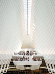The interior of a modern architectural structure features a high, vaulted ceiling with sleek, white rib-like beams that converge towards a central skylight. A group of people gather on multiple levels connected by stairways and platforms, creating a sense of activity within the expansive space. The design is futuristic and emphasizes symmetry and light.