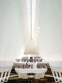 The interior of a modern architectural structure features a high, vaulted ceiling with sleek, white rib-like beams that converge towards a central skylight. A group of people gather on multiple levels connected by stairways and platforms, creating a sense of activity within the expansive space. The design is futuristic and emphasizes symmetry and light.