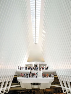 The interior of a modern architectural structure features a high, vaulted ceiling with sleek, white rib-like beams that converge towards a central skylight. A group of people gather on multiple levels connected by stairways and platforms, creating a sense of activity within the expansive space. The design is futuristic and emphasizes symmetry and light.