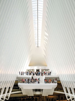The interior of a modern architectural structure features a high, vaulted ceiling with sleek, white rib-like beams that converge towards a central skylight. A group of people gather on multiple levels connected by stairways and platforms, creating a sense of activity within the expansive space. The design is futuristic and emphasizes symmetry and light.