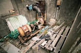 An abandoned industrial area with old, rusted machinery and scattered debris. A large pipe and electrical equipment are present, suggesting a past function in manufacturing or processing. The scene includes weathered wooden planks, a worn-down wall, and overgrown vegetation, contributing to a sense of decay.
