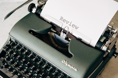 A vintage dark green typewriter with a sheet of paper loaded, displaying the word 'Review'. The typewriter is set against a light background, showcasing its classic keys and mechanical details.