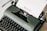 A vintage dark green typewriter with a sheet of paper loaded, displaying the word 'Review'. The typewriter is set against a light background, showcasing its classic keys and mechanical details.