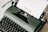 A vintage dark green typewriter with a sheet of paper loaded, displaying the word 'Review'. The typewriter is set against a light background, showcasing its classic keys and mechanical details.