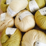 Several skeins of yarn arranged closely together, each labeled with product details. The yarn colors include variations of cream and olive green, and the labels are in black text on white paper.