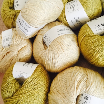 Several skeins of yarn arranged closely together, each labeled with product details. The yarn colors include variations of cream and olive green, and the labels are in black text on white paper.