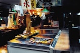 An indoor marketplace setting with various tools and utensils arranged on a metal counter. Overhead lights with green shades are illuminating the area, and a single white glove is suspended from the ceiling. Chinese characters appear on hanging signs. In the background, various items and a brightly lit stall add to the bustling atmosphere.