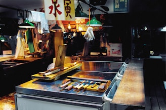 An indoor marketplace setting with various tools and utensils arranged on a metal counter. Overhead lights with green shades are illuminating the area, and a single white glove is suspended from the ceiling. Chinese characters appear on hanging signs. In the background, various items and a brightly lit stall add to the bustling atmosphere.