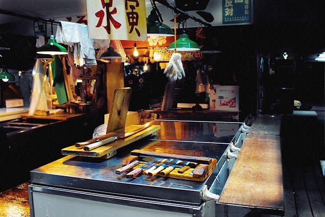 An indoor marketplace setting with various tools and utensils arranged on a metal counter. Overhead lights with green shades are illuminating the area, and a single white glove is suspended from the ceiling. Chinese characters appear on hanging signs. In the background, various items and a brightly lit stall add to the bustling atmosphere.