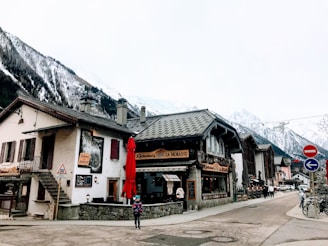 A quaint village scene featuring traditional Alpine-style buildings with steep roofs, surrounded by snow-capped mountains. There is a small café or restaurant with a red umbrella and a person walking by wearing winter clothes. The area seems quiet, with some people in the background. Road signs and bicycles are visible along the street.