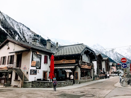 A quaint village scene featuring traditional Alpine-style buildings with steep roofs, surrounded by snow-capped mountains. There is a small café or restaurant with a red umbrella and a person walking by wearing winter clothes. The area seems quiet, with some people in the background. Road signs and bicycles are visible along the street.