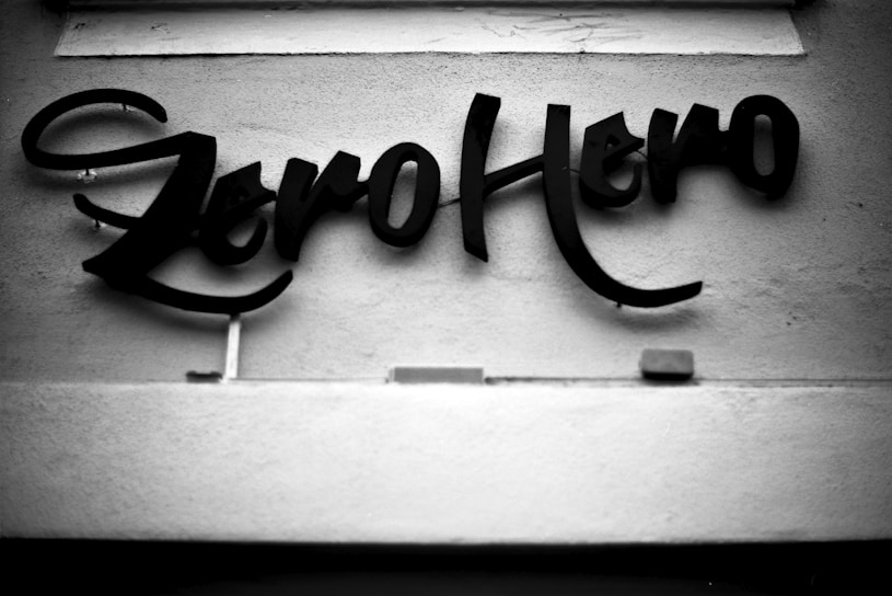 Elegant, cursive lettering reads 'Zero Hero' against a textured wall. The black text stands out starkly against the smooth, light-colored background.