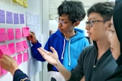 A group of people engaged in a collaborative discussion by a whiteboard covered in colorful sticky notes. The participants appear to be focused and are gesturing towards the notes, indicating a brainstorming session or planning activity. Various notes have handwritten text, suggesting ideas or tasks.