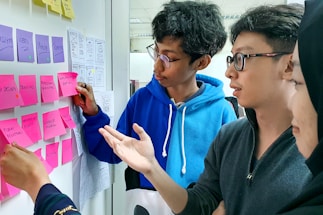 A group of people engaged in a collaborative discussion by a whiteboard covered in colorful sticky notes. The participants appear to be focused and are gesturing towards the notes, indicating a brainstorming session or planning activity. Various notes have handwritten text, suggesting ideas or tasks.