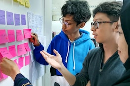 A group of people engaged in a collaborative discussion by a whiteboard covered in colorful sticky notes. The participants appear to be focused and are gesturing towards the notes, indicating a brainstorming session or planning activity. Various notes have handwritten text, suggesting ideas or tasks.