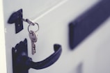 A close-up of a door with a key inserted in the lock. The key is attached to a keyring, and the door handle is visible below. The door appears to be painted in a light color, contrasting with the darker handle and lock.