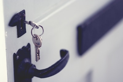 A close-up of a door with a key inserted in the lock. The key is attached to a keyring, and the door handle is visible below. The door appears to be painted in a light color, contrasting with the darker handle and lock.